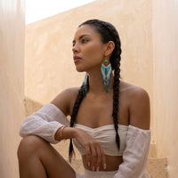 Woman with braided hair wearing large turquoise earrings against a beige wall.