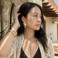 Woman wearing gold jewelry outdoors near a stone wall. Molten Flame Statement Earrings - The Songbird Collection
