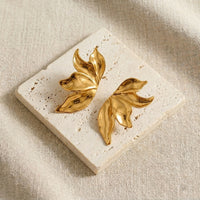 Gold leaf-shaped earrings on a textured stone surface with a beige fabric background.  Molten Flame Statement Earrings - The Songbird Collection 
