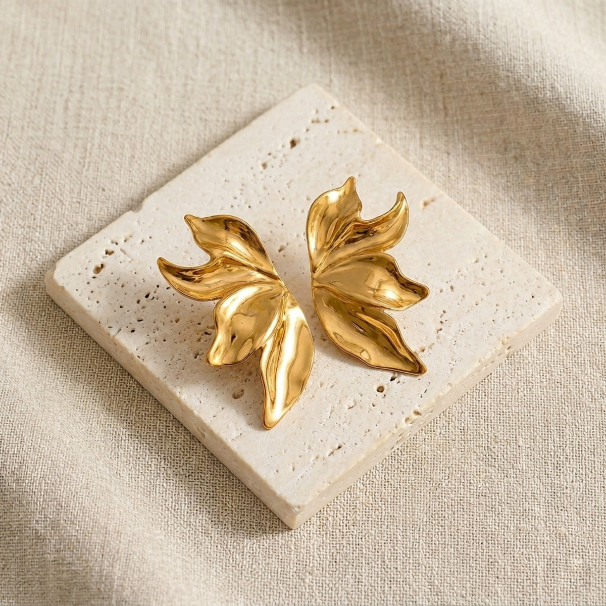 Gold leaf-shaped earrings on a beige stone surface.  Molten Flame Statement Earrings - The Songbird Collection 