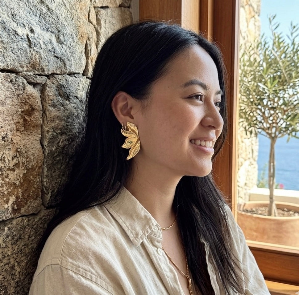 Woman with long dark hair wearing gold earrings, leaning against a stone wall with a scenic background.  Molten Flame Statement Earrings - The Songbird Collection