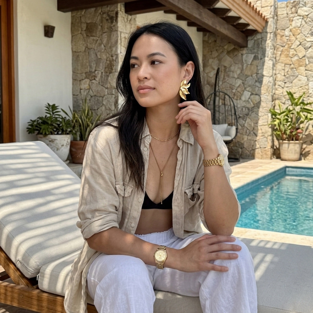 Woman sitting by a poolside with a stone wall and plants in the background.  Molten Flame Statement Earrings - The Songbird Collection