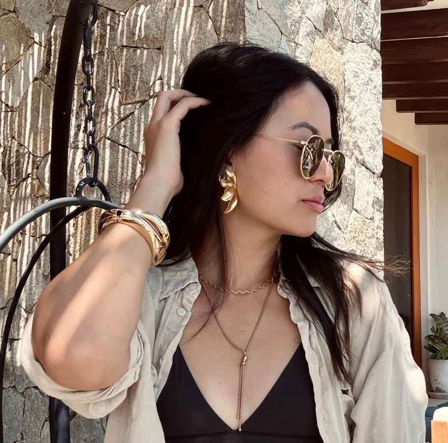 Woman wearing sunglasses and gold jewelry outdoors against a stone wall.  Molten Flame Statement Earrings - The Songbird Collection