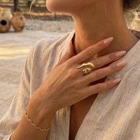 Close-up of a person wearing a gold ring and bracelet, with a blurred outdoor background.  Molten Gold Statement Ring - The Songbird Collection