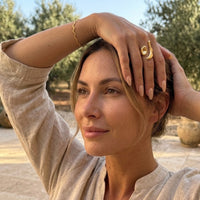 Woman with hand on head outdoors, wearing a gold ring. Molten Gold Statement Ring , Aura Ripple Bracelet - The Songbird Collection