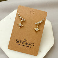 A pair of gold-colored ear crawlers with cubic zirconia and a star drop pendant, displayed on a fabric surface.