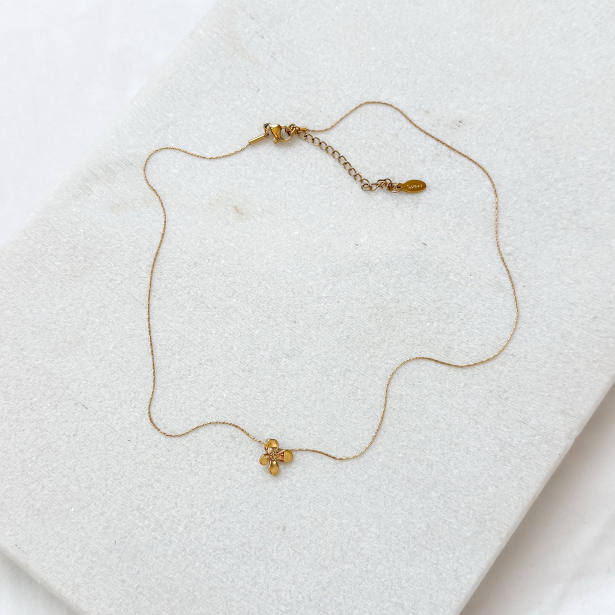 A delicate gold chain necklace with a small gold flower charm, displayed on a textured white surface.  Petite Petal Necklace - The Songbird Collection