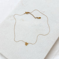 A delicate gold chain necklace with a small gold flower charm, displayed on a textured white surface.  Petite Petal Necklace - The Songbird Collection