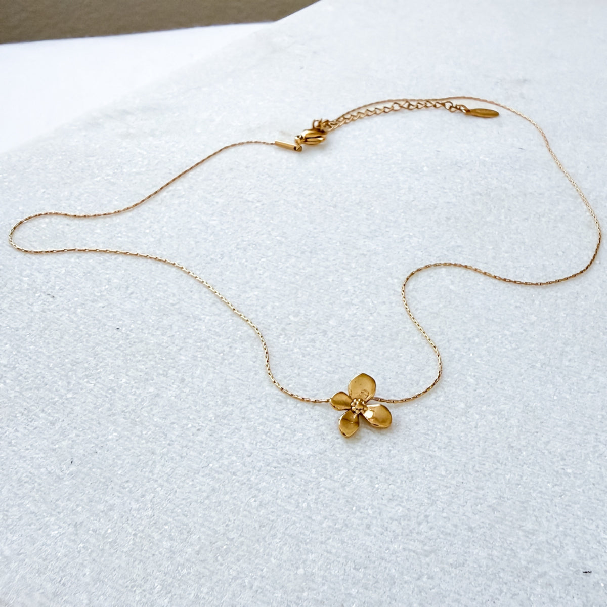A delicate gold chain necklace featuring a small, textured gold flower pendant, displayed on a textured white surface.  Petite Petal Necklace - The Songbird Collection