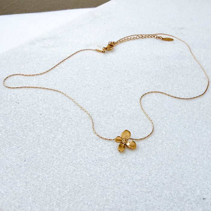 A delicate gold chain necklace featuring a small, textured gold flower pendant, displayed on a textured white surface.  Petite Petal Necklace - The Songbird Collection