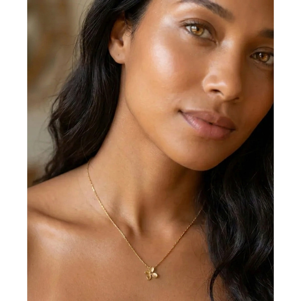 A close-up of a woman wearing a delicate gold chain necklace with a small gold flower or clover pendant, shown against her neck and upper chest.  Petite Petal Necklace - The Songbird Collection