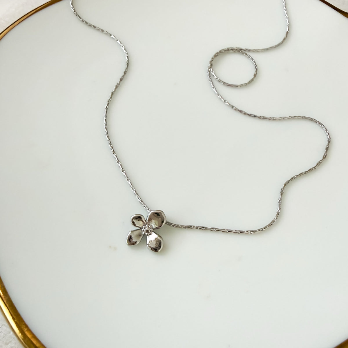 A delicate silver chain necklace with a small textured silver flower pendant, displayed on a white ceramic plate with a gold rim.  Petite Petal Necklace - The Songbird Collection