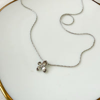 A delicate silver chain necklace with a small textured silver flower pendant, displayed on a white ceramic plate with a gold rim.  Petite Petal Necklace - The Songbird Collection
