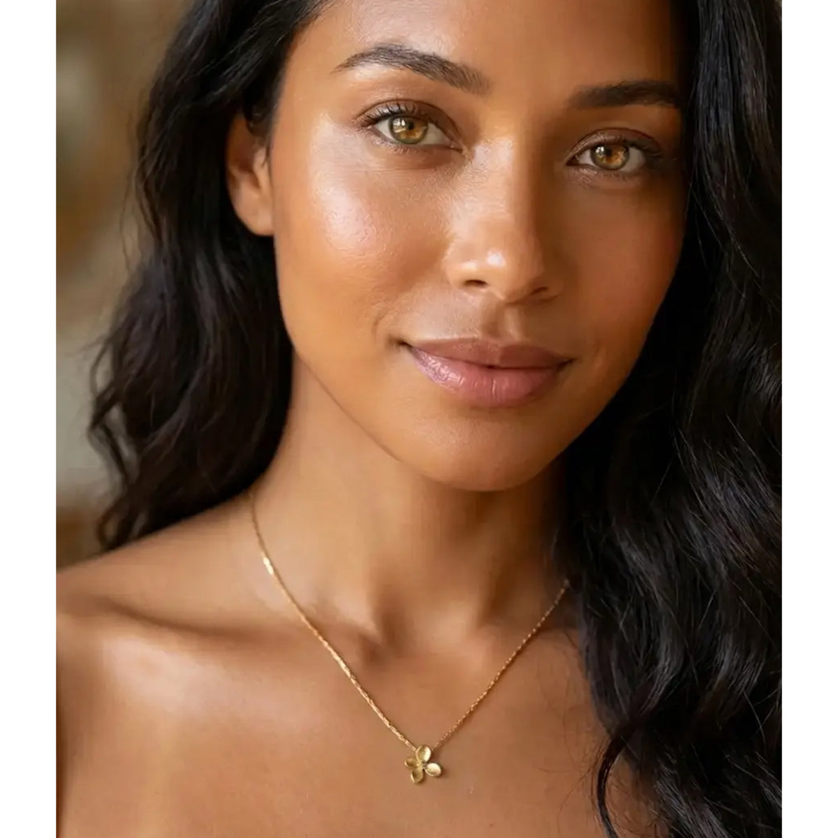 A close-up portrait of a woman with long dark wavy hair wearing a delicate gold chain necklace with a small gold flower pendant, set against a softly blurred warm background.  Petite Petal Necklace - The Songbird Collection