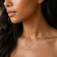 A close-up portrait of a woman with long dark wavy hair wearing a delicate gold chain necklace with a small gold flower pendant, set against a softly blurred warm background.  Petite Petal Necklace - The Songbird Collection