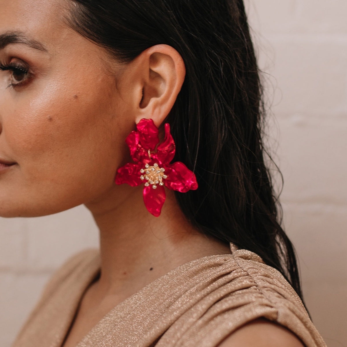 A person wearing a beige top and Rue Flower Earrings in red, which are large, pearlescent floral earrings with rhinestone centers.