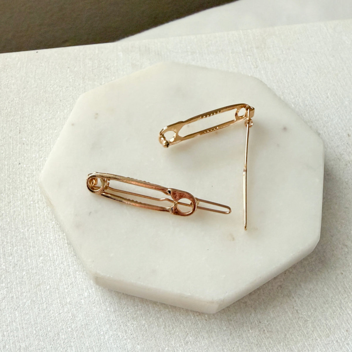 Two gold safety-pin style earrings displayed on a white octagonal stone tray over a light fabric surface.  Safety Pin Hair Pin Set - The Songbird Collection