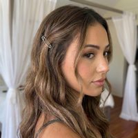 A woman with long, wavy brown hair with blonde highlights, wearing a silver safety pin hair clip on the side of her hair, shown in a softly lit indoor setting with white curtains.  Safety Pin Hair Pin Set - The Songbird Collection