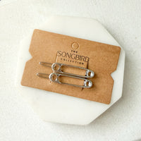 Two silver safety-pin style hair pins displayed on a brown card labeled 'THE SONGBIRD COLLECTION,' placed on a white octagonal stand against a light neutral background.