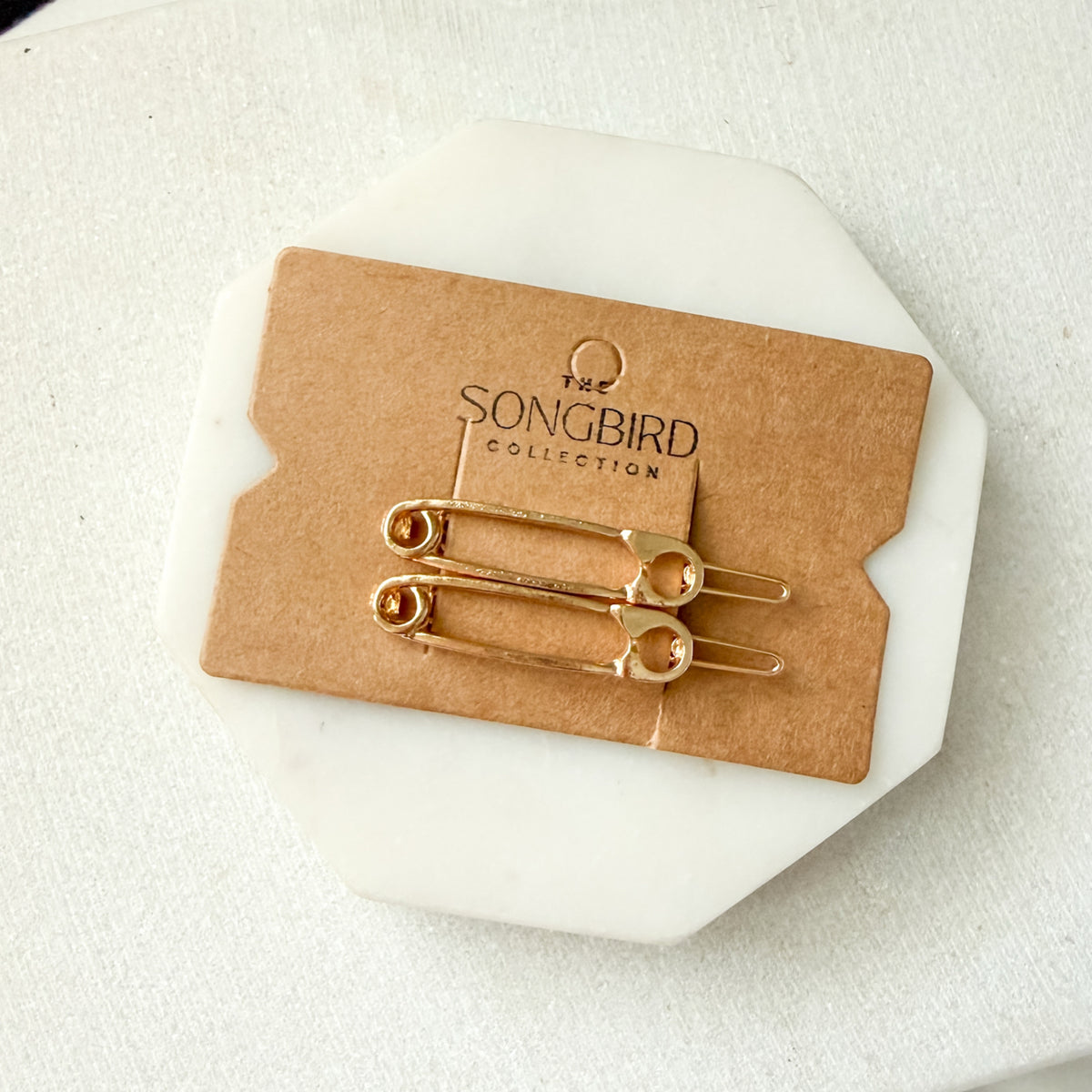 Two gold safety-pin style hair pins attached to a brown kraft display card, placed on a white octagonal stand over a light fabric background. The card is branded 'THE SONGBIRD COLLECTION'.