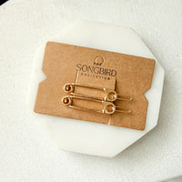 Two gold safety-pin style hair pins attached to a brown kraft display card, placed on a white octagonal stand over a light fabric background. The card is branded 'THE SONGBIRD COLLECTION'.