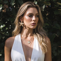 Woman wearing a white top with a starfish earring and necklace, standing against a leafy background.  Starfish_Statement_Earrings_-_The_Songbird_Collection_