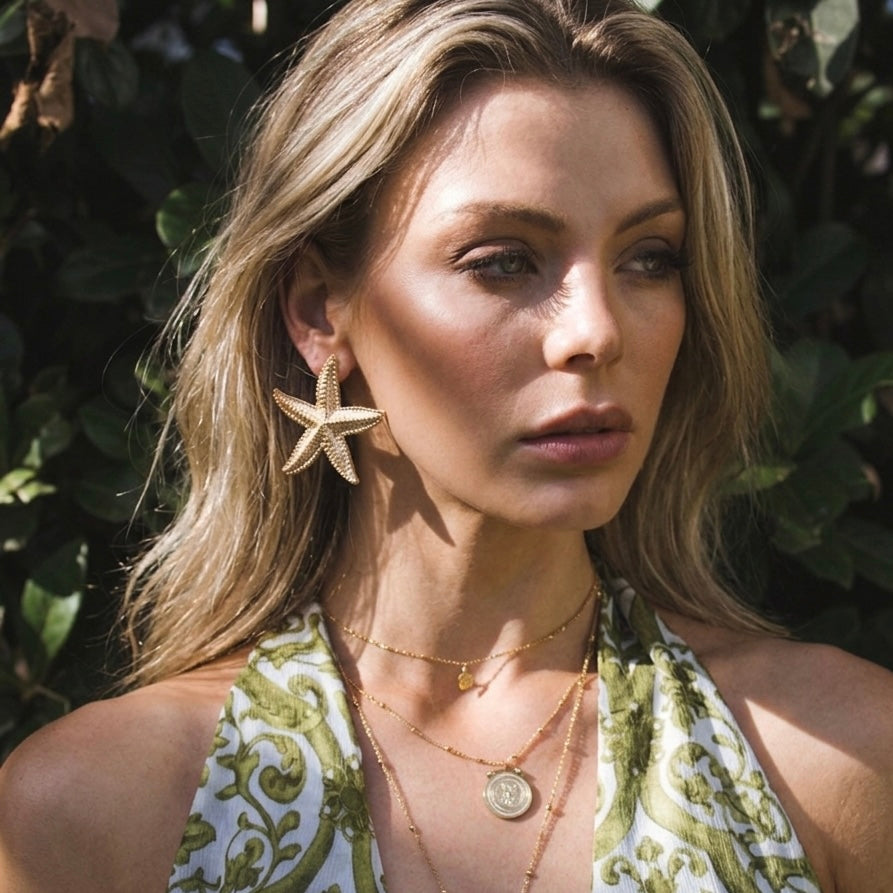 Woman wearing starfish earrings and necklaces with a green and white patterned top, surrounded by foliage.  Starfish_Statement_Earrings_-_The_Songbird_Collection_