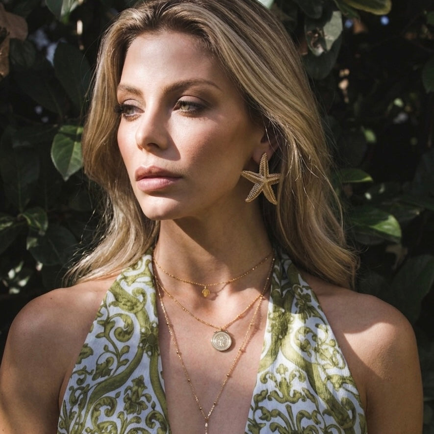 Woman wearing a green and white patterned top with layered necklaces and starfish earrings against a leafy background.  Starfish_Statement_Earrings_-_The_Songbird_Collection_