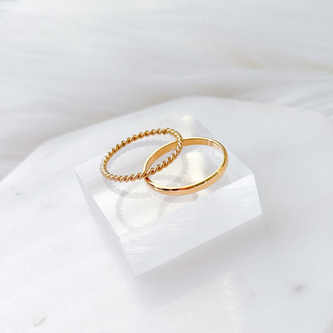 Two gold-plated stainless steel rings on a white marble surface, one with a smooth band and the other with a twisted chain band.