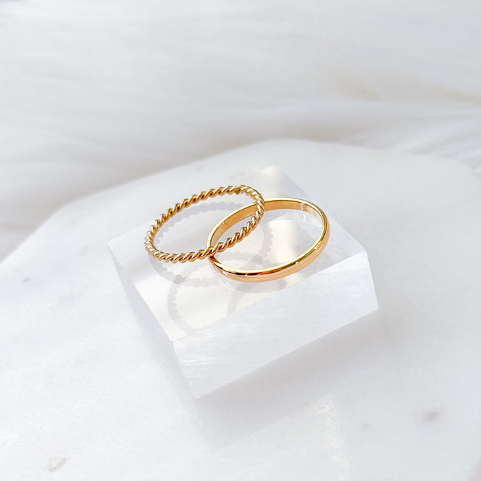 Two gold-plated stainless steel rings on a white marble surface, one with a smooth band and the other with a twisted chain band.