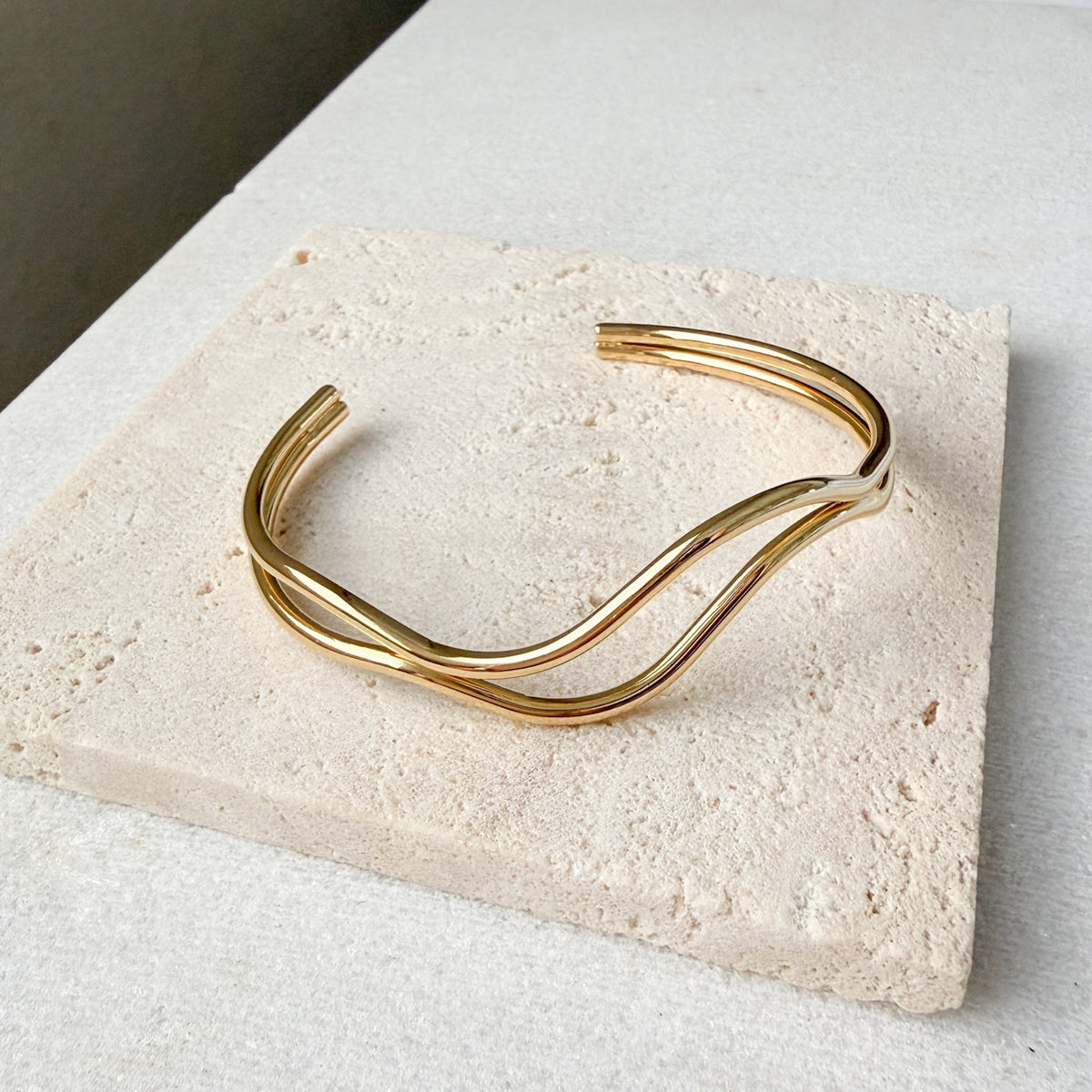Gold bracelet  double wavy lines on a textured stone surface. The Contour Cuff Bracelet - The Songbird Collection