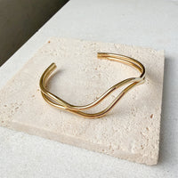 Gold bracelet  double wavy lines on a textured stone surface. The Contour Cuff Bracelet - The Songbird Collection