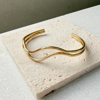 Gold bracelet  double wavy lines on a textured stone surface. The Contour Cuff Bracelet - The Songbird Collection