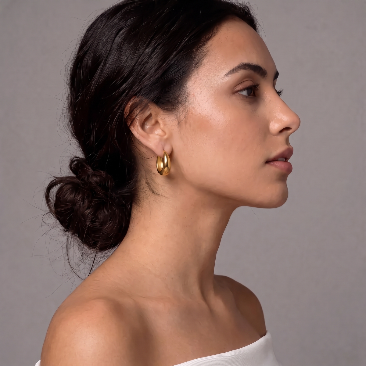 A woman in profile wearing a pair of small gold hoop earrings against a plain gray background.  The Ever Teardrop Hoop Earrings - The Songbird Collection