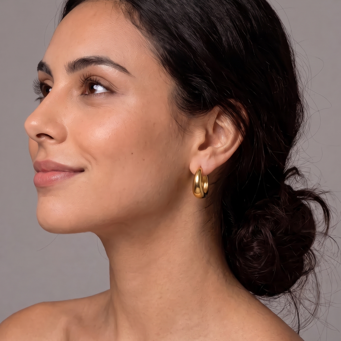 A close-up side profile of a woman with dark hair in a low bun, wearing a small gold hoop earring against a plain gray background.  The Ever Teardrop Hoop Earrings - The Songbird Collection