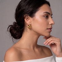A close-up side profile of a woman with dark hair in a low bun, wearing a pair of gold hoop earrings, against a neutral gray background.  The Ever Teardrop Hoop Earrings - The Songbird Collection