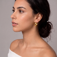A close-up side profile of a woman wearing a small gold hoop earring against a plain gray studio background.  The Ever Teardrop Hoop Earrings - The Songbird Collection