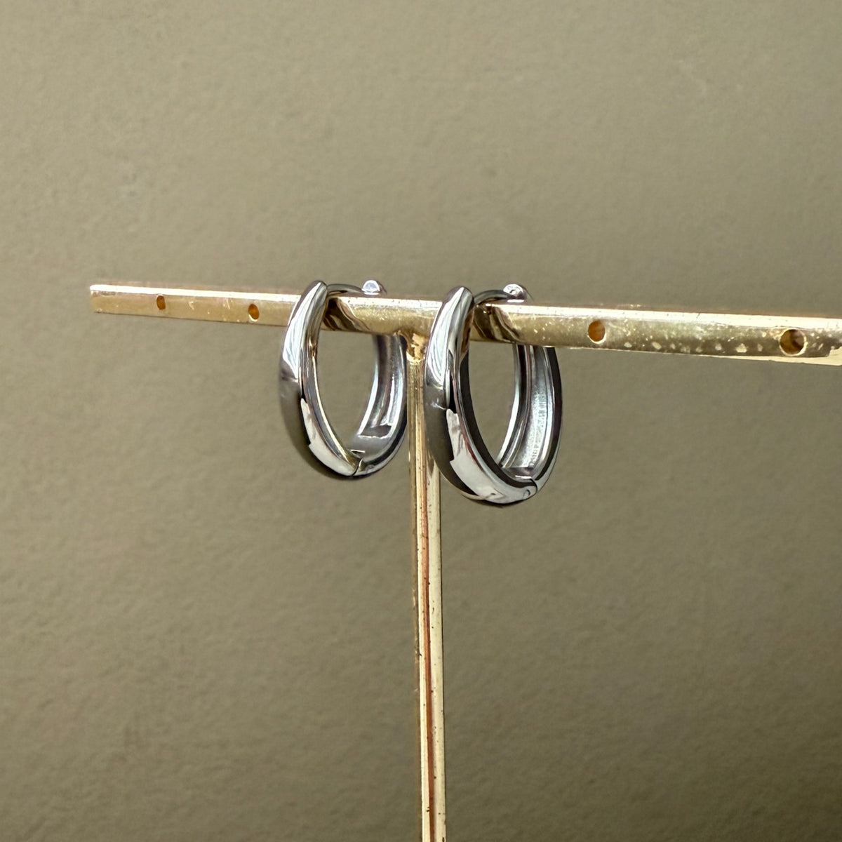 Two silver  oblong teardrop hoop earrings on a gold stand against a beige background