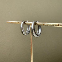 Two silver  oblong teardrop hoop earrings on a gold stand against a beige background
