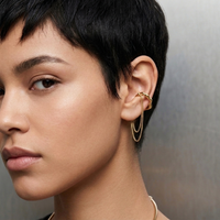 A close-up of a model with short dark hair wearing a delicate gold ear cuff with two hanging chain drops on her ear.   The Flow Ear Cuff - The Songbird Collection