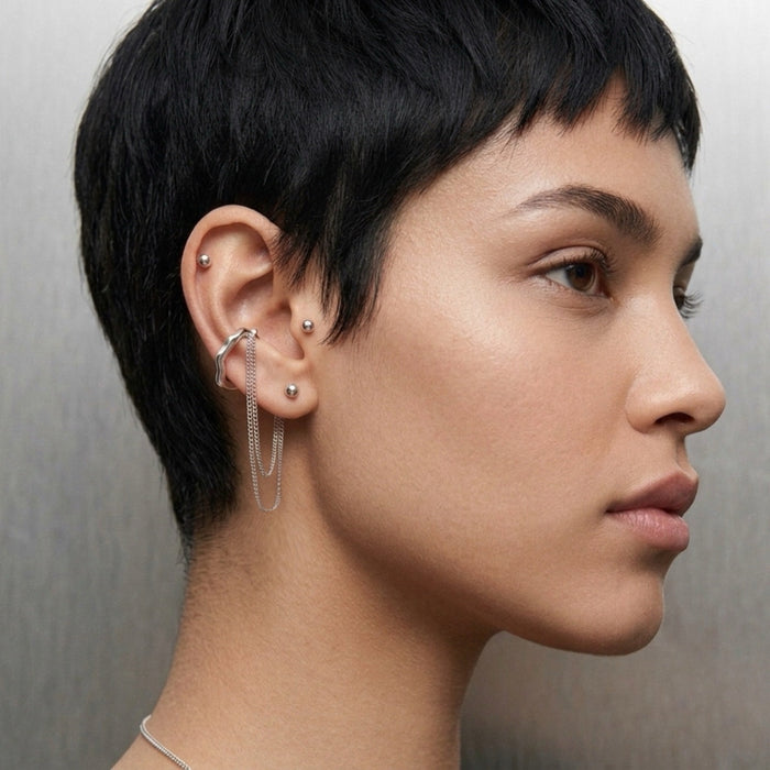 A close-up side profile of a model with a short dark pixie cut, showcasing a silver ear cuff with two hanging chains and multiple silver stud and hoop earrings on her ear, against a neutral gray background.  The Flow Ear Cuff - The Songbird Collection