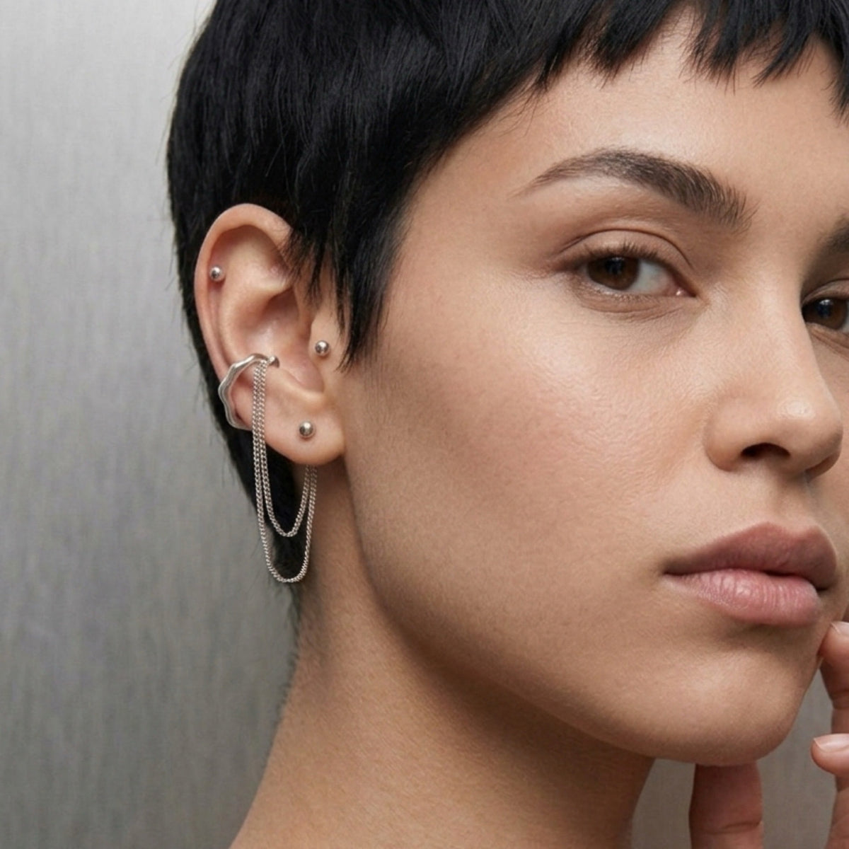 Close-up of a person with short dark hair wearing multiple silver ear piercings, including a large chain hoop piercing through the lobe and small studs on other ear locations, against a neutral gray background.  The Flow Ear Cuff - The Songbird Collection