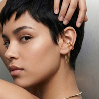 Close-up of a model with short dark hair wearing a delicate gold ear cuff with chain drapes, against a soft gray background.  The Flow Ear Cuff - The Songbird Collection