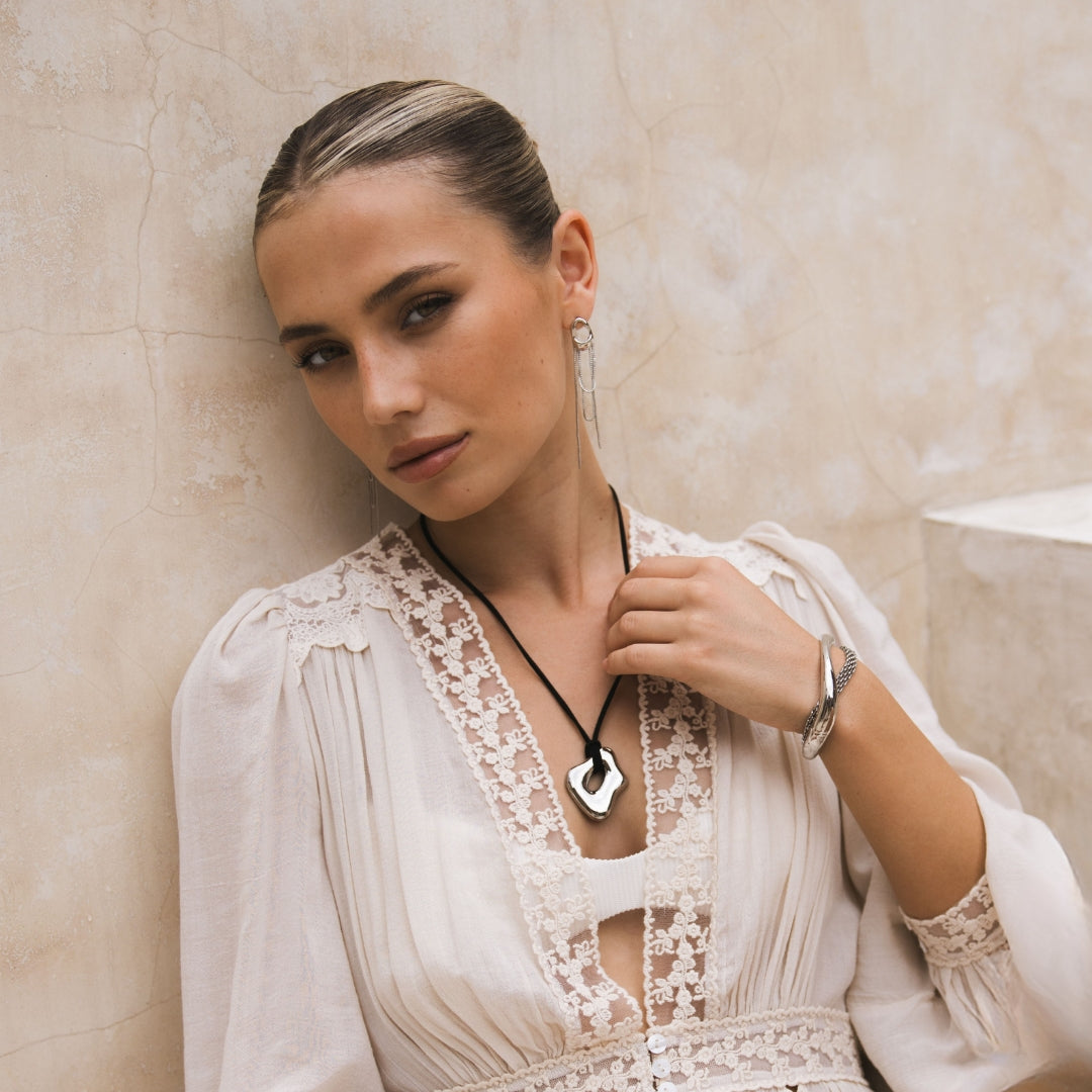 Woman wearing a white lace dress with draped chain earrings, and a black rope pendant necklace against a beige wall