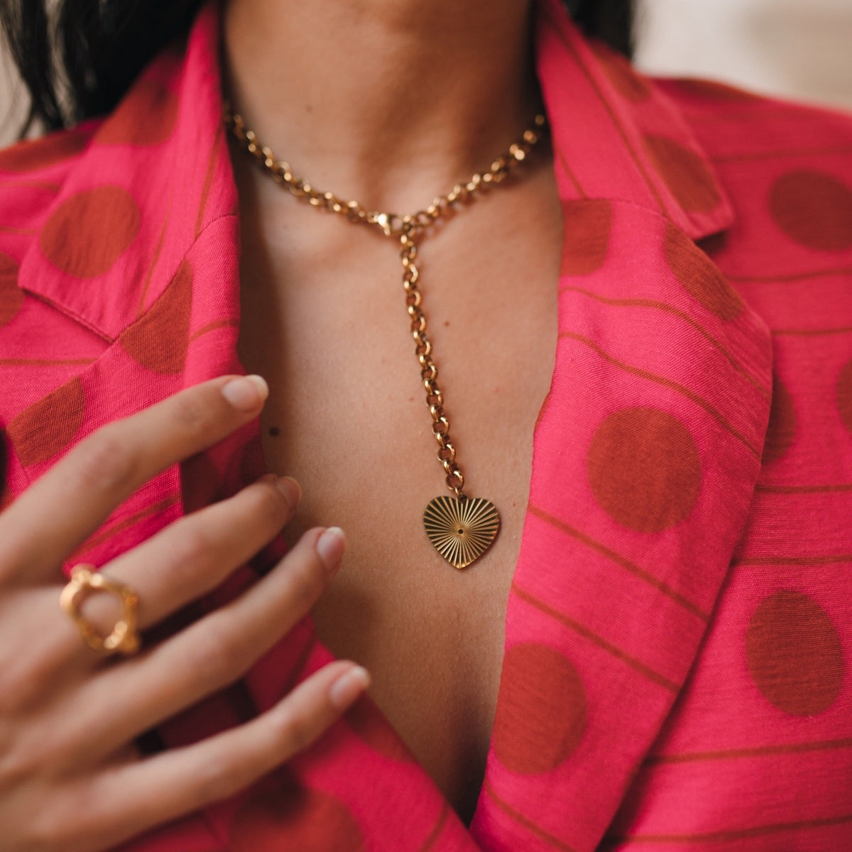 A close-up image of a person wearing a Treasured Heart Necklace with a gold-plated heart pendant and chain, paired with a pink garment with polka dots. Treasured Heart Necklace - The Songbird Collection