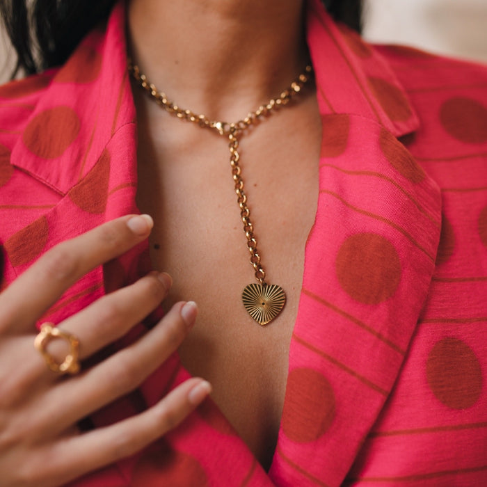 A close-up image of a person wearing a Treasured Heart Necklace with a gold-plated heart pendant and chain, paired with a pink garment with polka dots. Treasured Heart Necklace - The Songbird Collection