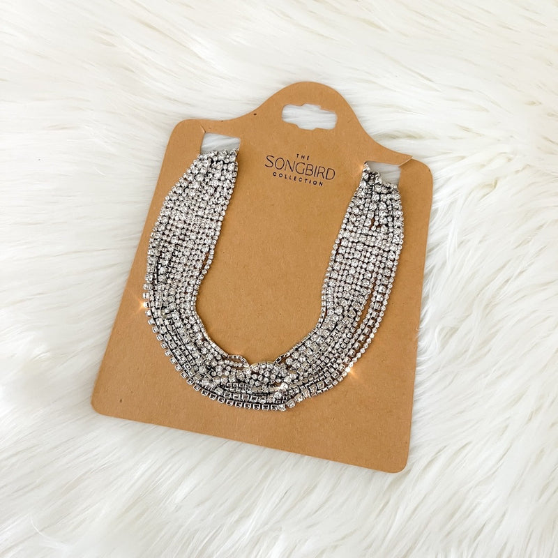 A rhinestone necklace displayed on a beige card against a white fluffy background.