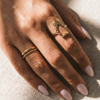 Close-up of a hand wearing two gold rings on a textured beige background gothic_throne_ring - Tessa Ring Set- The Songbird Collection