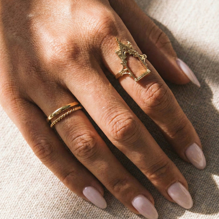 Close-up of a hand wearing two gold rings on a textured beige background gothic_throne_ring - Tessa Ring Set- The Songbird Collection