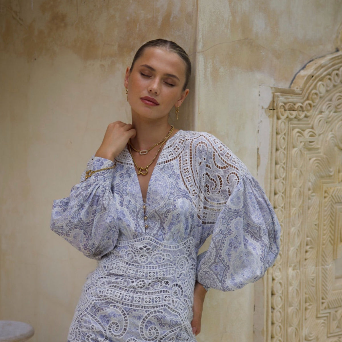 A woman in a white, lace-patterned dress with balloon sleeves stands against a textured beige wall, eyes closed and hand on her neck, wearing The Songbird Collection’s Icon Link Necklace and gold jewelry, her hair pulled back.